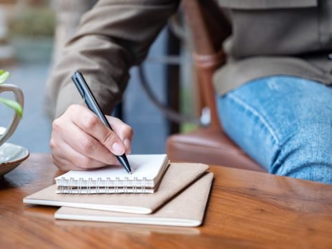Closeup image of a woman's hand writing on blank notebook with coffee cup on wooden table